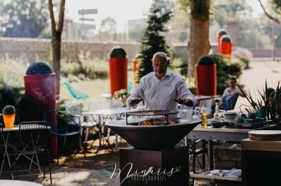 Un chef cuisine sur un brasero dans un jardin, entouré de tables et de végétation.