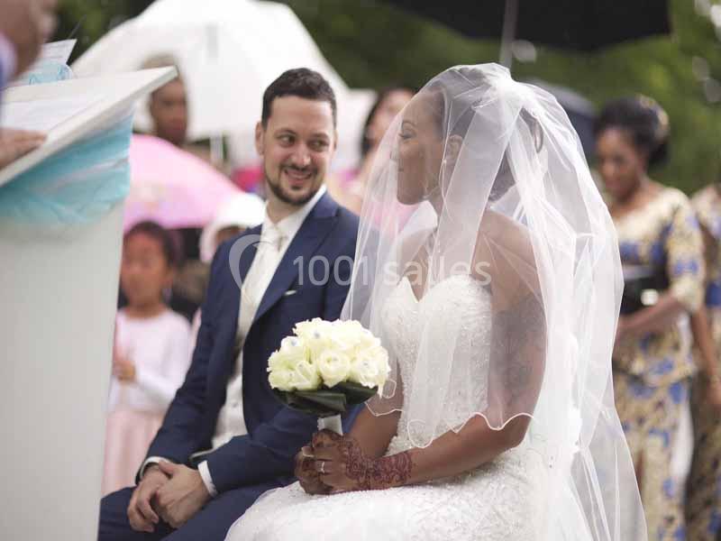 Un couple assis lors d'une cérémonie de mariage en extérieur, la mariée tenant un bouquet de fleurs blanches.