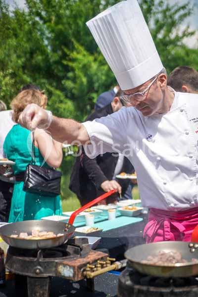 Un chef en tenue blanche cuisine en extérieur, ajoutant des ingrédients dans une poêle sur un plan de travail.