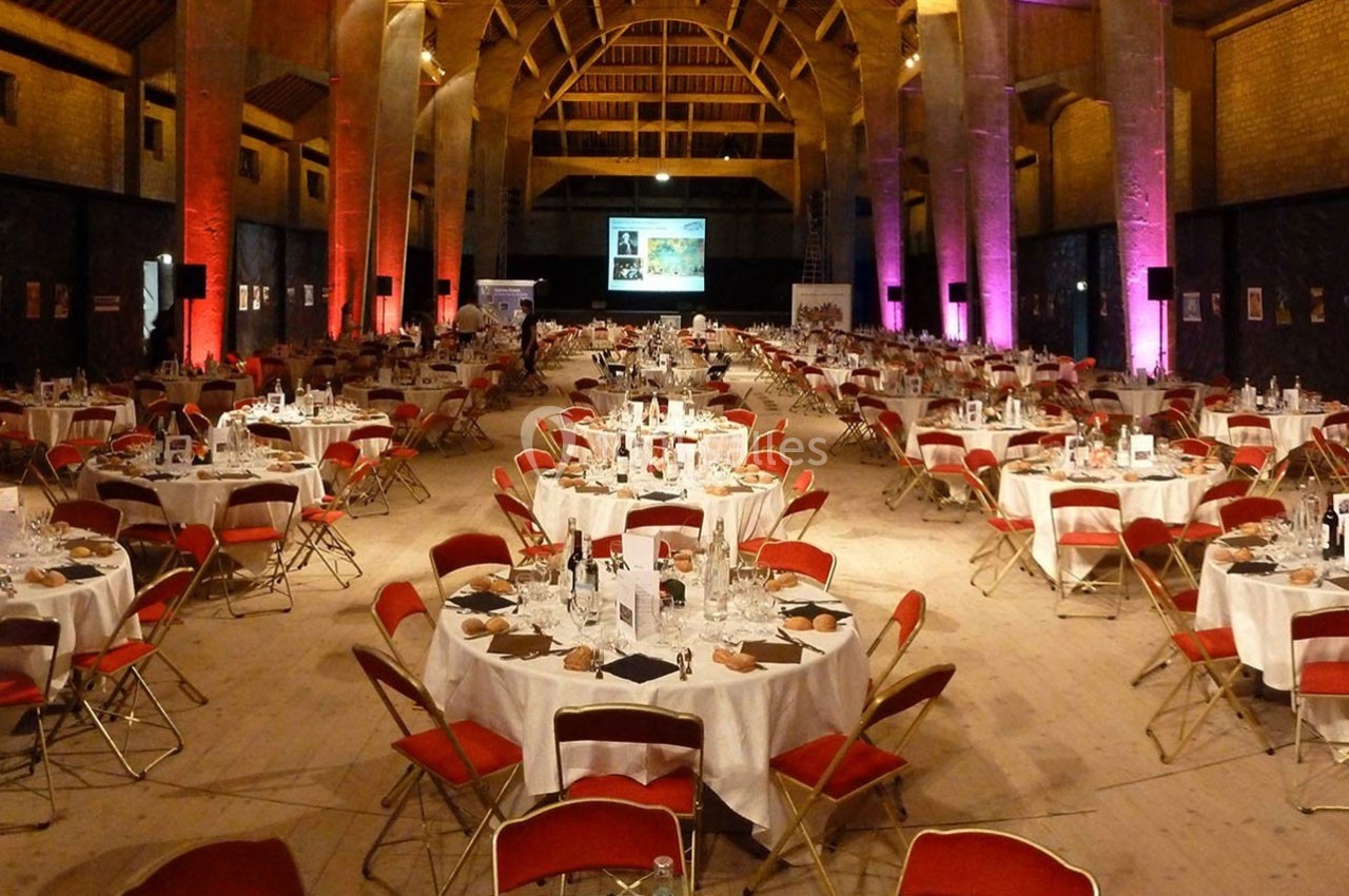 Salle spacieuse aménagée pour un dîner, avec des tables rondes dressées et des chaises rouges, éclairage chaleureux.