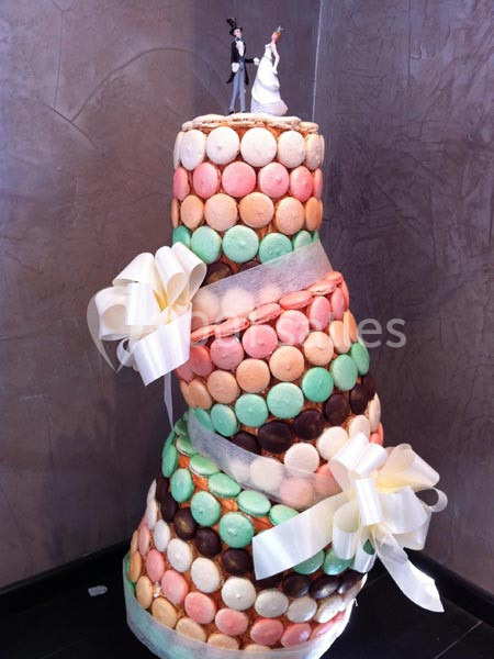 Pièce montée inclinée décorée de macarons colorés, rubans blancs et figurines de mariés au sommet.