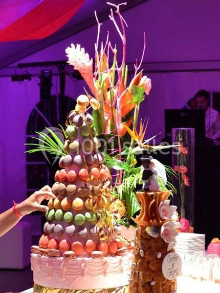 Tour de macarons colorés décorée de fleurs tropicales, avec une pièce montée en arrière-plan dans un cadre festif.