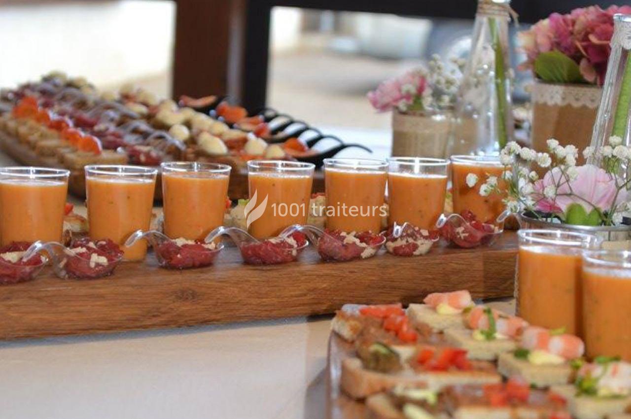 Buffet avec verrines de soupe orange, cuillères de charcuterie et toasts garnis, décoré de fleurs en arrière-plan.