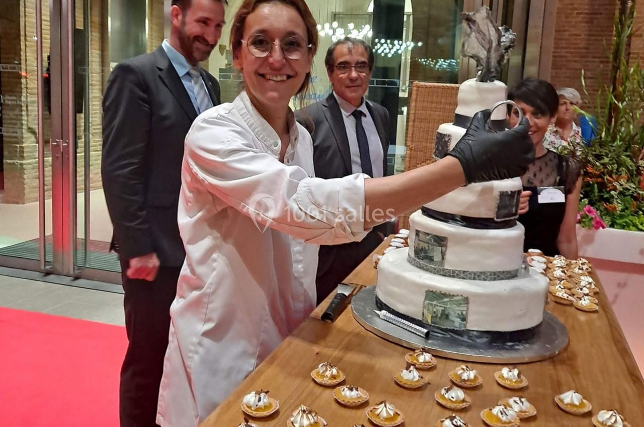 Une pâtissière souriante découpe un gâteau décoré, entourée de convives lors d'un événement festif.