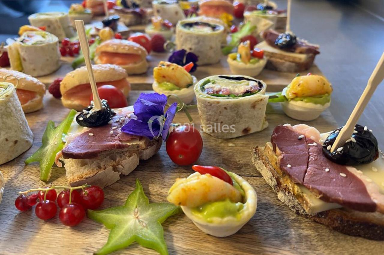 Plateau de canapés variés avec fruits, légumes, viandes et fleurs comestibles, présenté sur une planche en bois.