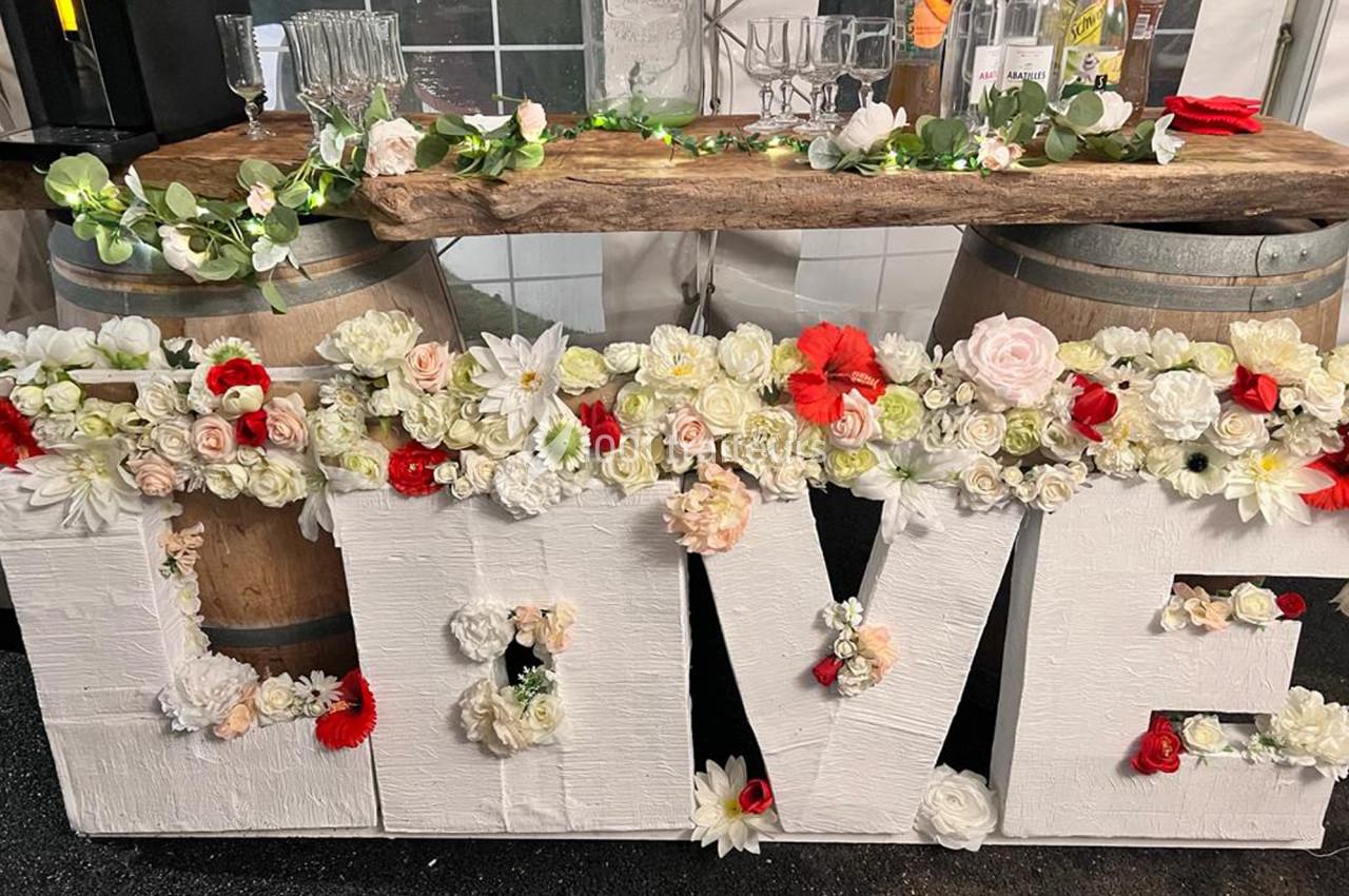 Décoration de mariage avec le mot ’LOVE’ en lettres blanches ornées de fleurs, devant une table en bois sur tonneaux.