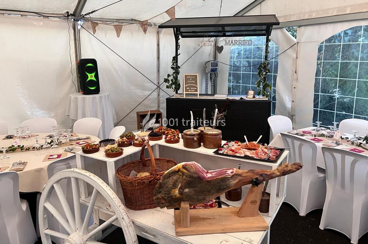 Buffet de mariage sous une tente, avec tables dressées, un jambon sur support et divers plats disposés sur une charrette.