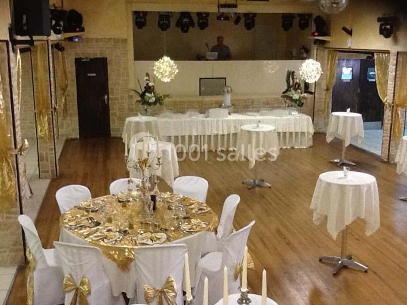 Salle de réception décorée avec des tables rondes et hautes, nappes blanches et accents dorés, prête pour un événement.