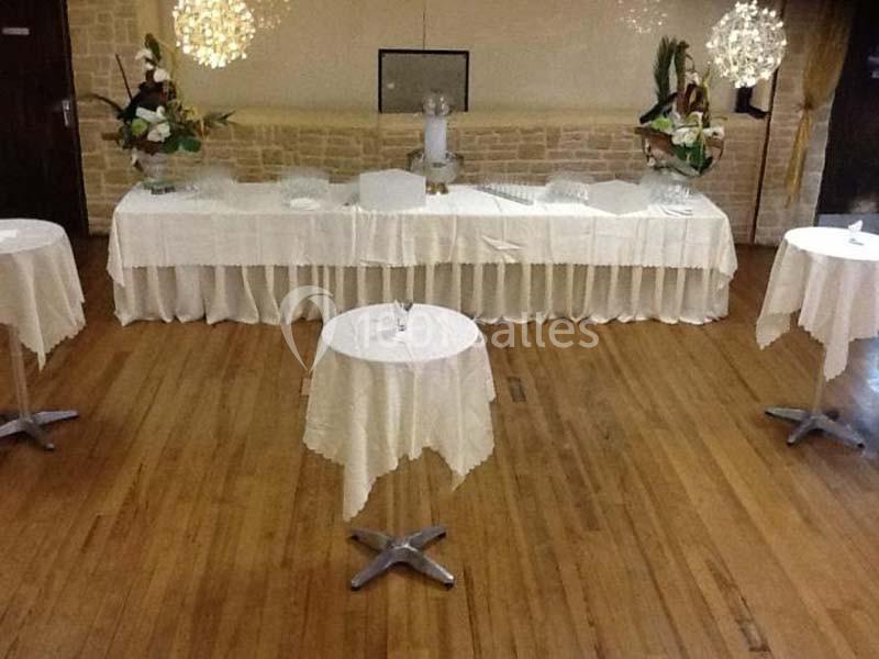 Salle décorée avec des tables rondes et un buffet recouvert de nappes blanches, sur un sol en parquet.