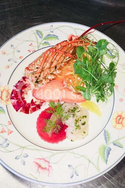 Assiette gastronomique composée de homard, tranches de poisson, légumes frais et herbes, présentée sur une vaisselle fleurie.