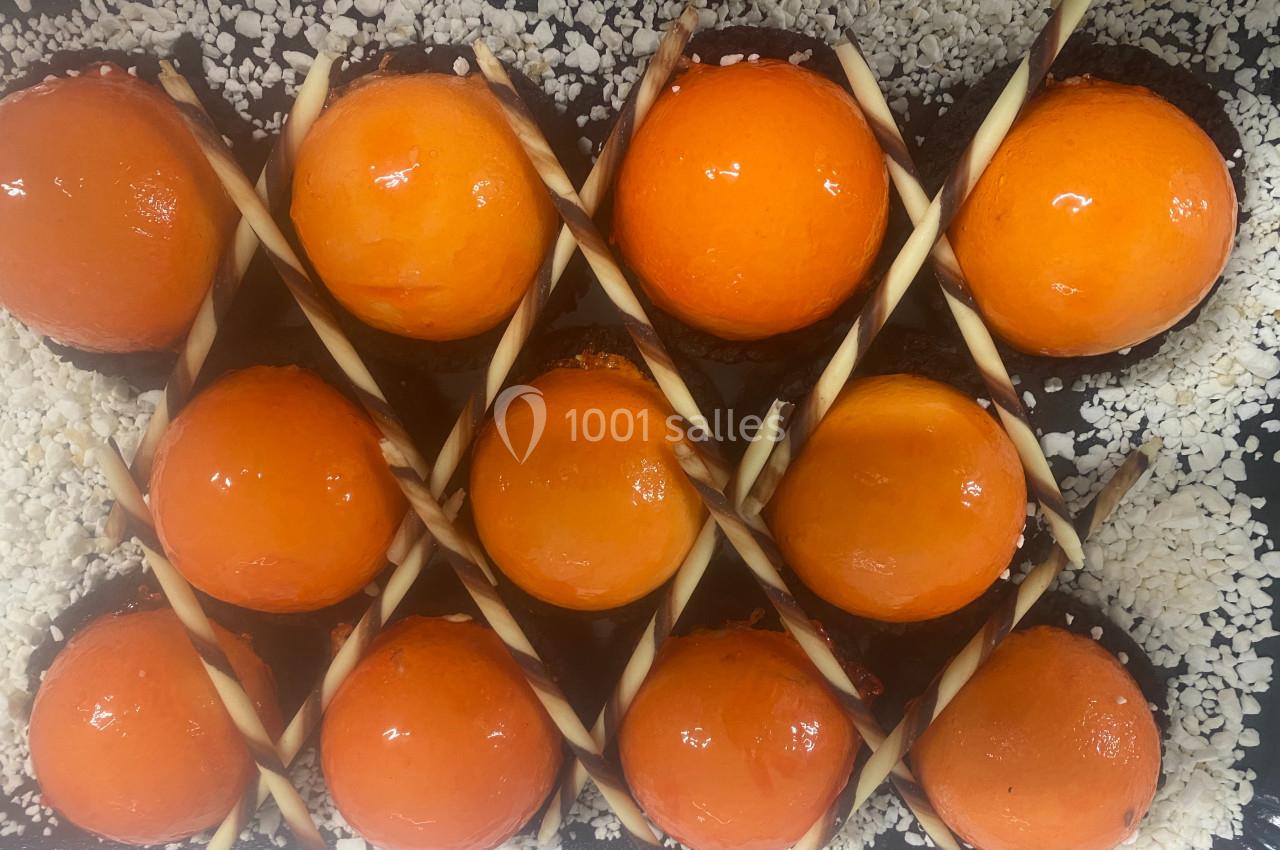 Dessert composé de sphères orange brillantes sur une base chocolatée, décoré de bâtonnets marbrés et de copeaux blancs.