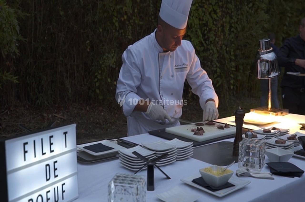 Un chef en tenue blanche découpe du filet de bœuf sur une table dressée en extérieur, éclairée par une lampe.