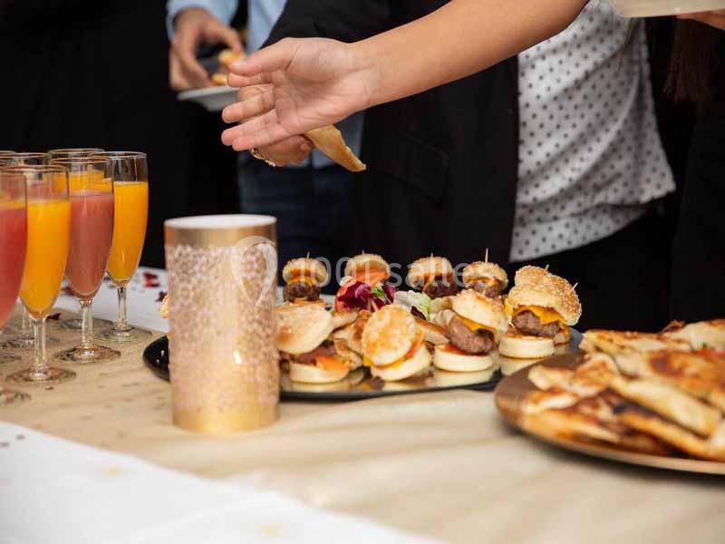 Une main prend un amuse-bouche sur une table garnie de mini-burgers, cocktails et autres plats lors d'un buffet.