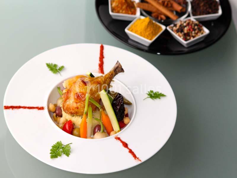 Plat de couscous garni de légumes, poulet rôti et épices, présenté dans une assiette blanche sur une table en verre.