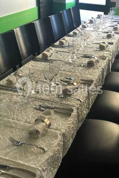 Table dressée avec une nappe grise à motifs, couverts, verres et serviettes pliées, entourée de chaises noires.