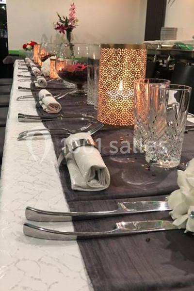 Table dressée avec nappe blanche et chemin de table gris, couverts alignés, serviettes roulées et bougie décorative allumée.