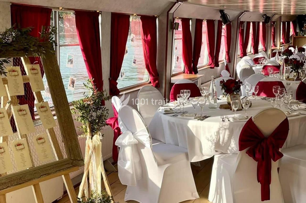 Salle de réception décorée avec des nappes blanches, des rubans rouges et un plan de table sur chevalet.