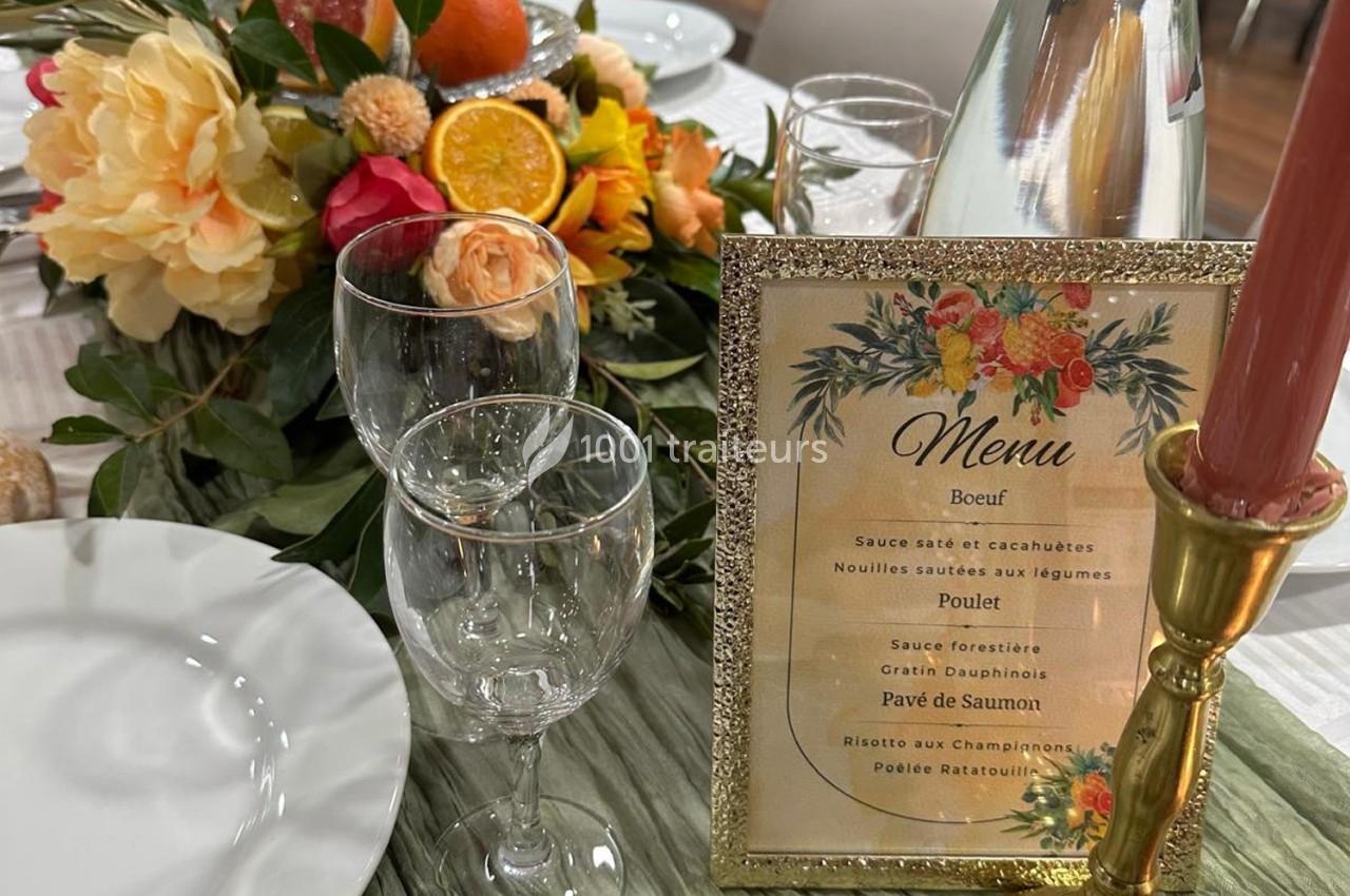Menu encadré sur une table dressée avec des verres, une bougie, des assiettes et une décoration florale colorée.