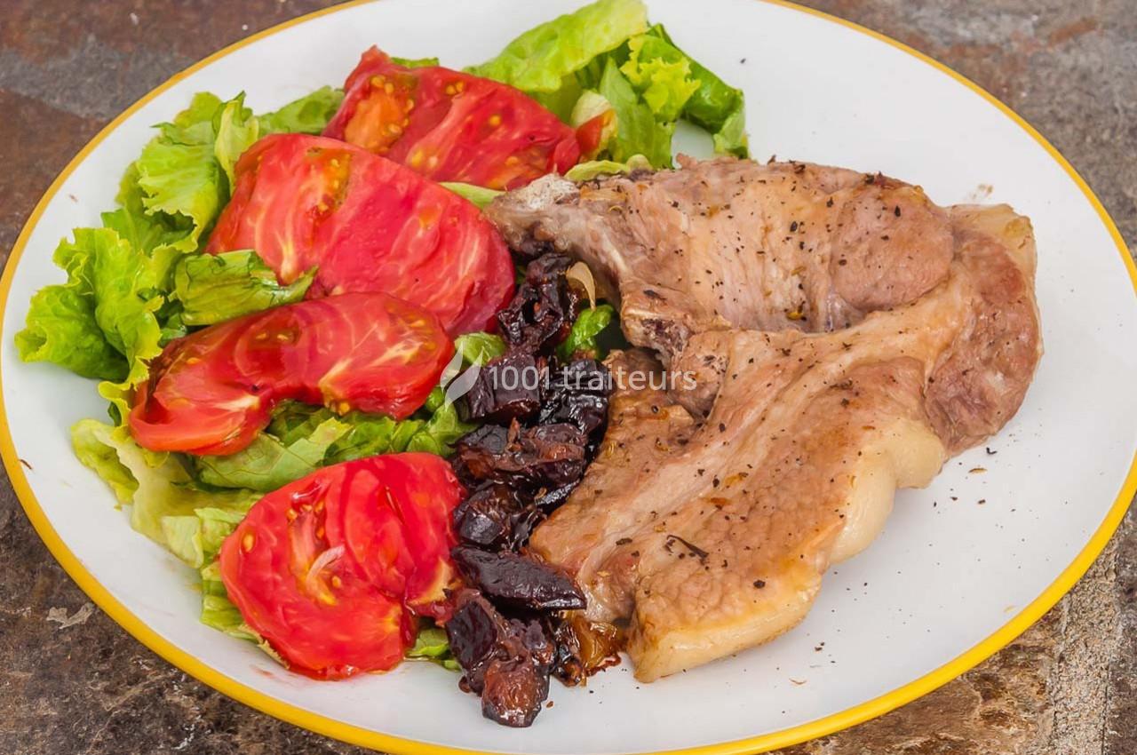 Assiette contenant une côte de porc grillée, des tranches de tomate, de la salade verte et des morceaux d'olives noires.