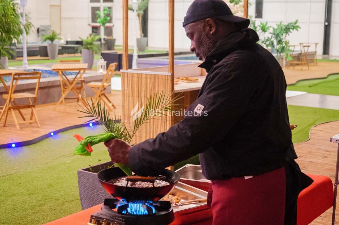 Un cuisinier prépare un plat dans une poêle sur un réchaud, dans un espace extérieur aménagé avec des plantes et des tables.