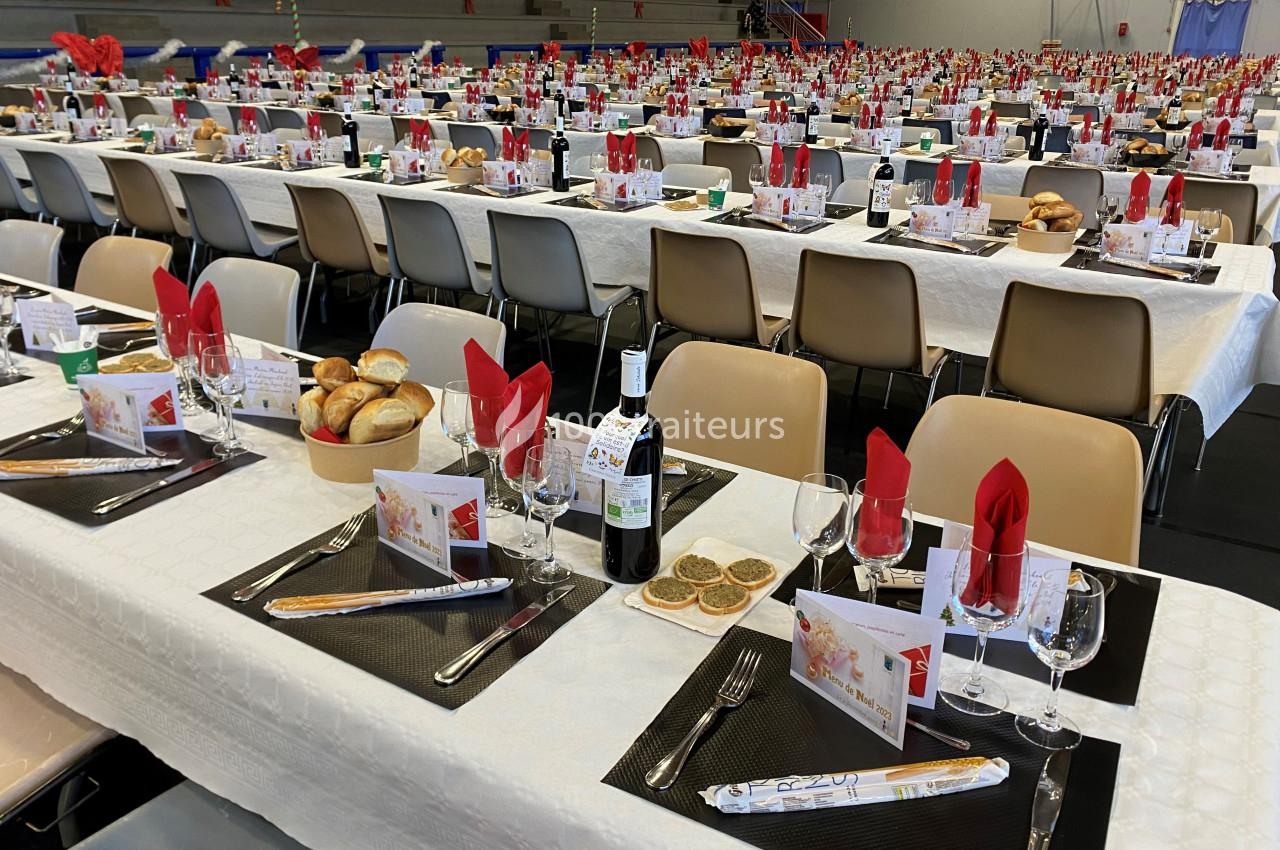 Tables dressées pour un grand repas, avec nappes blanches, serviettes rouges, couverts, pain et bouteilles de vin.