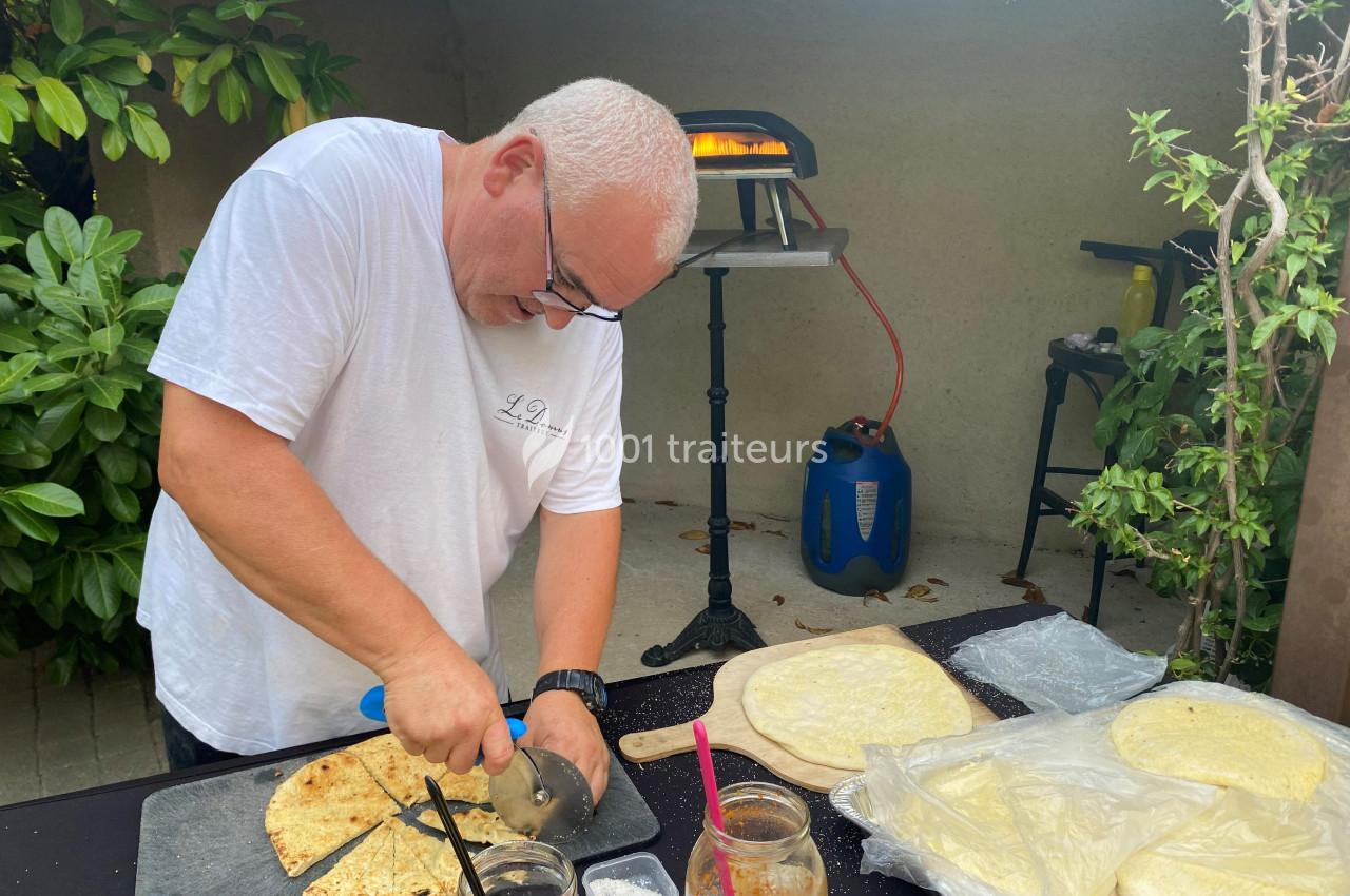 Un homme coupe une pizza cuite sur une table extérieure près d'un four à pizza, entouré de végétation.