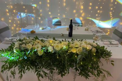 Centre de table floral avec roses et magnolias, bouteille de vin, numéro de table et décorations sur une nappe blanche.