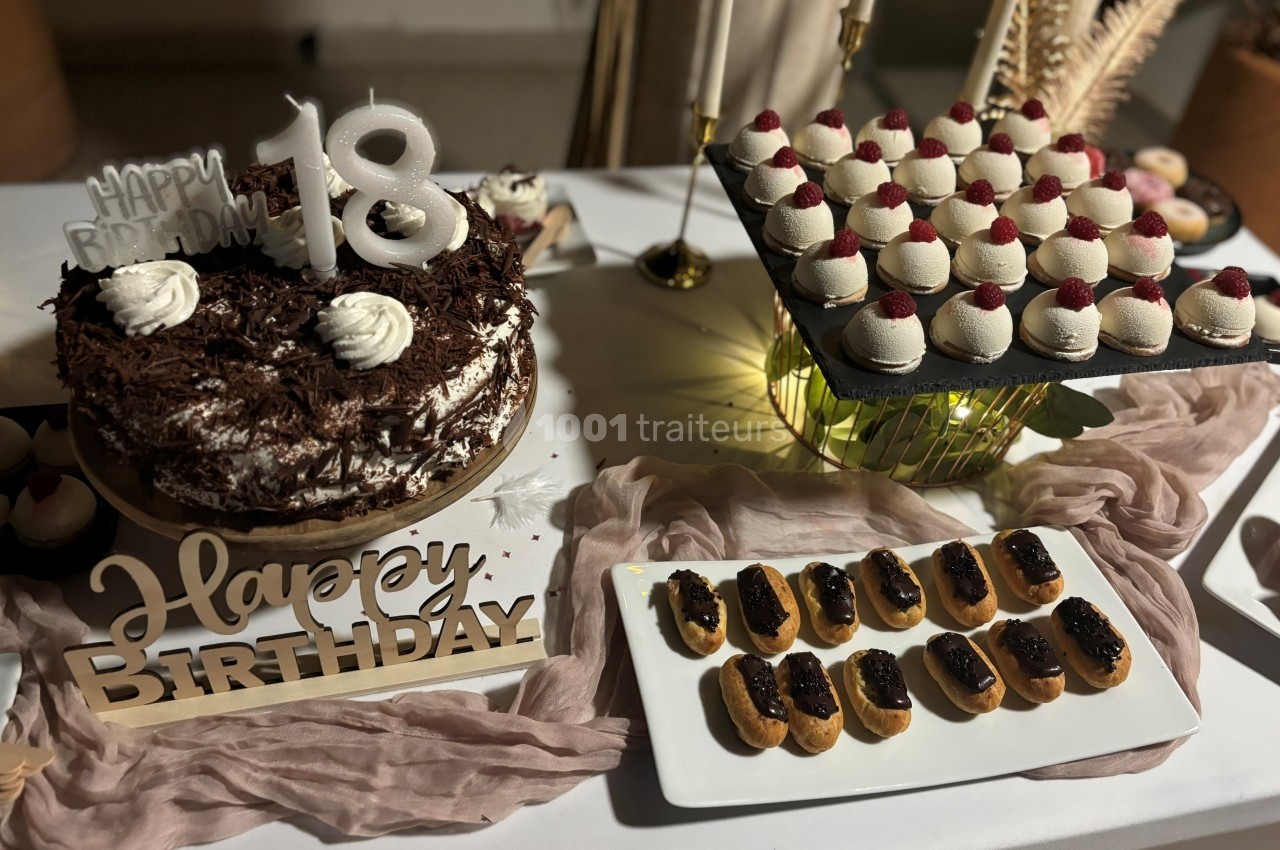 Table décorée avec un gâteau d'anniversaire, des éclairs au chocolat et des mignardises garnies de framboises.