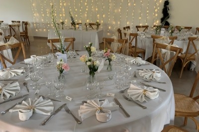 Centre de table floral avec roses et magnolias, bouteille de vin, numéro de table et décorations sur une nappe blanche.