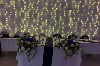 Centre de table floral avec roses et magnolias, bouteille de vin, numéro de table et décorations sur une nappe blanche.
