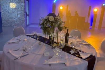Centre de table floral avec roses et magnolias, bouteille de vin, numéro de table et décorations sur une nappe blanche.