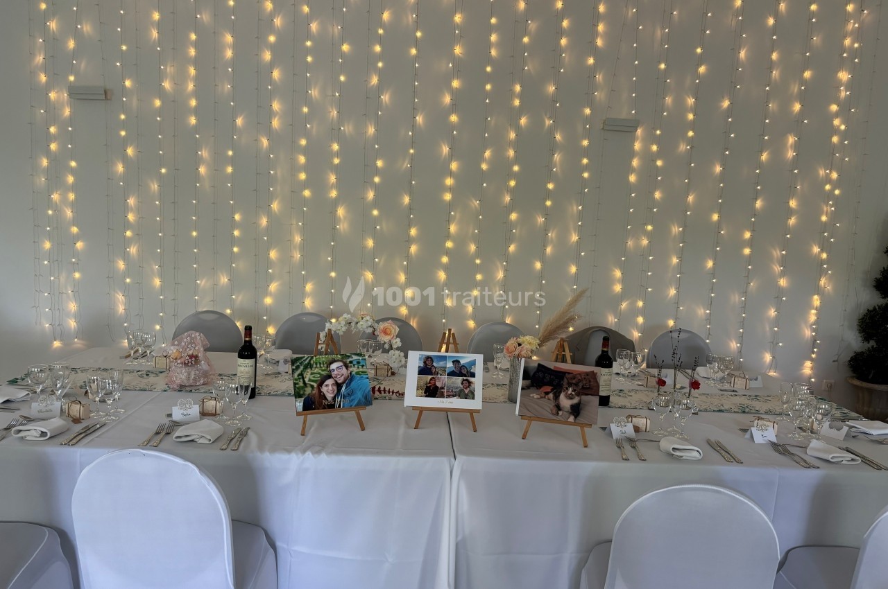 Table décorée pour un événement avec nappes blanches, guirlandes lumineuses en arrière-plan et photos sur chevalets.