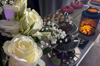 Centre de table floral avec roses et magnolias, bouteille de vin, numéro de table et décorations sur une nappe blanche.