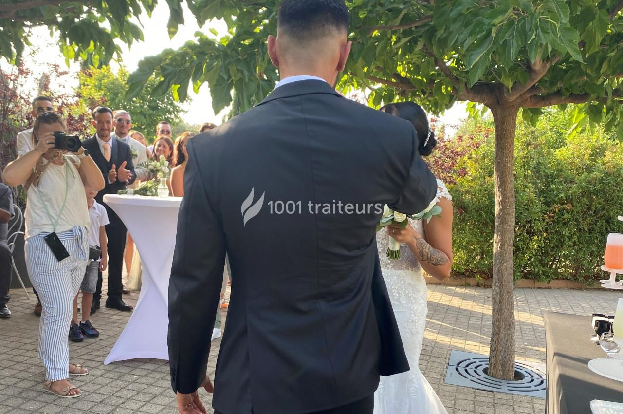 Un homme en costume sombre et une femme en robe de mariée avancent sous des arbres, entourés d'invités.