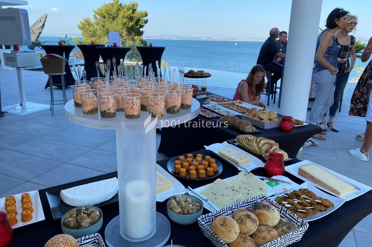 Buffet en extérieur avec vue sur la mer, comprenant pains, viennoiseries, fromages et verrines disposés sur des tables.