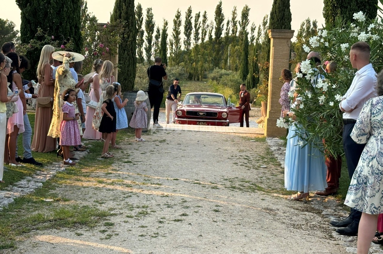 Une voiture ancienne rouge arrive sur une allée bordée d'invités en tenue élégante lors d'un événement en plein air.