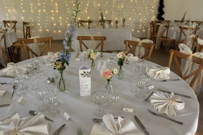 Centre de table floral avec roses et magnolias, bouteille de vin, numéro de table et décorations sur une nappe blanche.