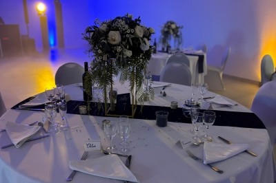 Centre de table floral avec roses et magnolias, bouteille de vin, numéro de table et décorations sur une nappe blanche.