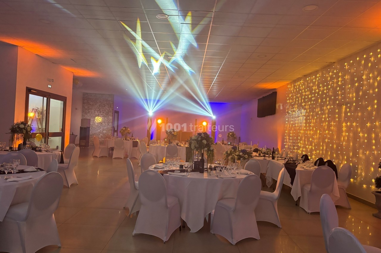 Salle de réception décorée avec des tables rondes, des chaises blanches, des lumières colorées et un mur illuminé.
