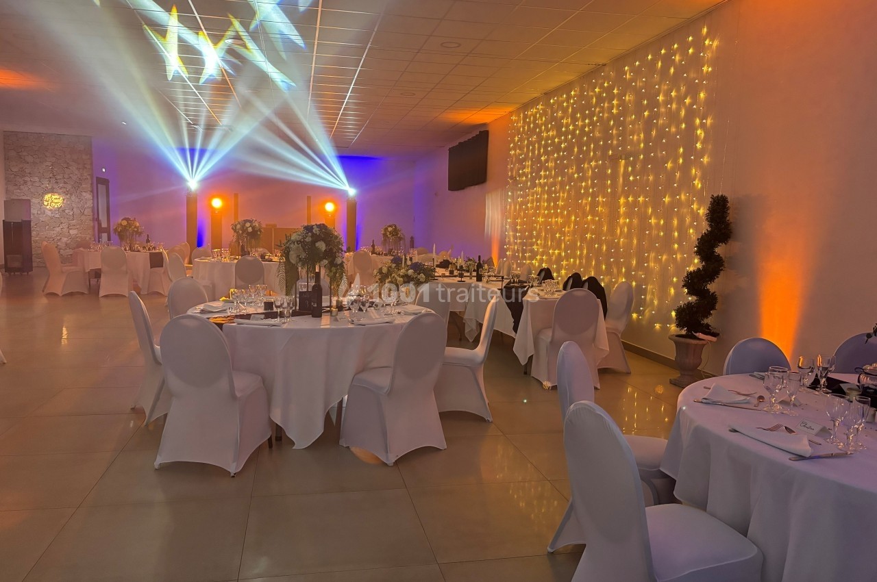 Salle de réception décorée avec des tables dressées, éclairages colorés et guirlandes lumineuses sur un mur.