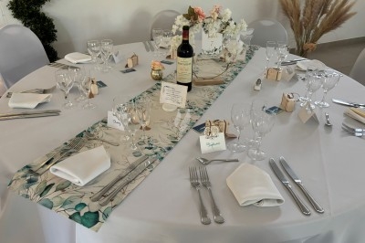 Centre de table floral avec roses et magnolias, bouteille de vin, numéro de table et décorations sur une nappe blanche.
