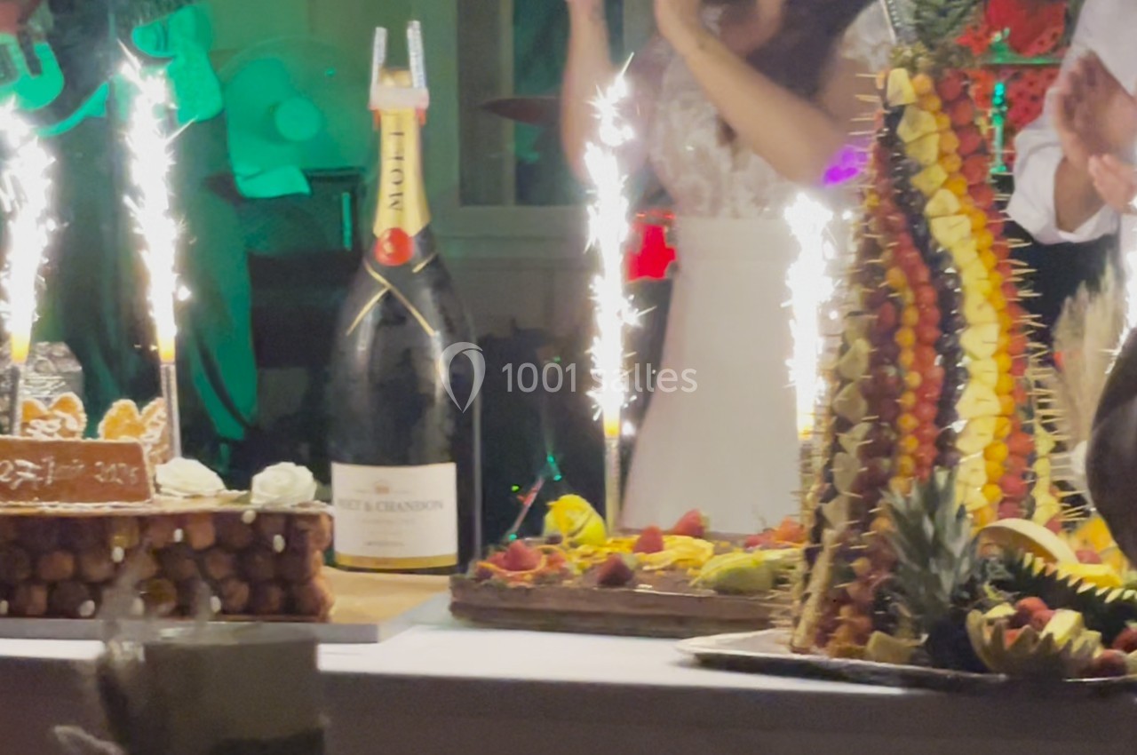 Table festive avec pièce montée de fruits, gâteau, bouteille de champagne et cierges scintillants allumés.