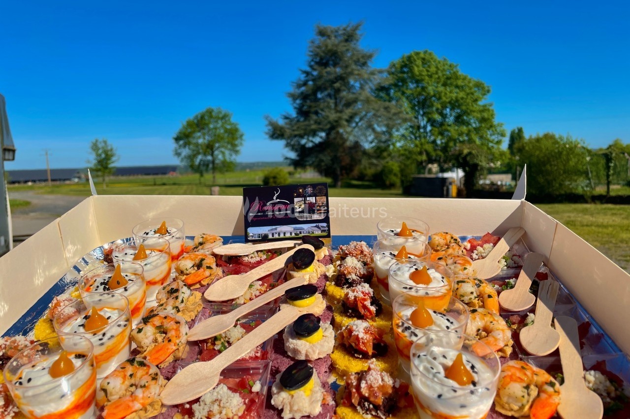Plateau de bouchées apéritives variées avec verrines, toasts et cuillères en bois, présenté en extérieur sous un ciel bleu.