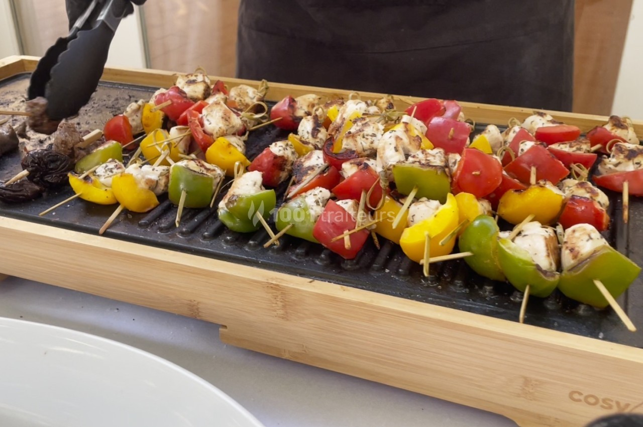 Brochettes de légumes et de poulet grillées sur une plancha en bois, avec des pinces tenant un morceau.