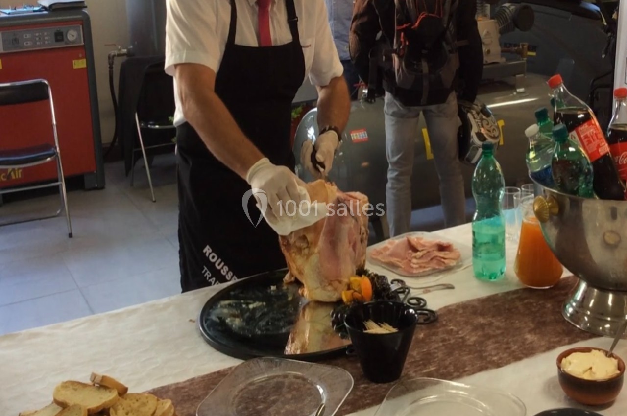 Un homme découpe un jambon sur un support, entouré de boissons et d'ustensiles sur une table.