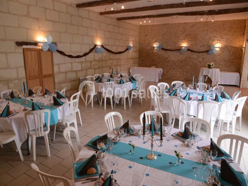 Salle décorée pour un événement avec des tables rondes dressées, nappes bleues et blanches, et chaises en plastique blanc.