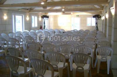 Salle avec des rangées de chaises en plastique blanc disposées face à une scène lumineuse dans un espace aux murs clairs.