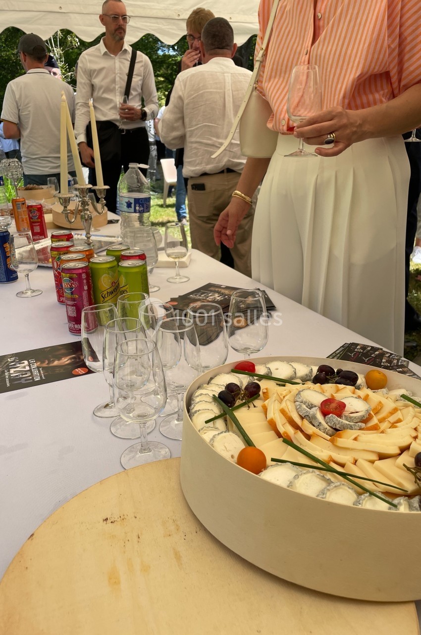 Plateau de fromages variés décoré de fruits, posé sur une table avec verres, boissons et convives en arrière-plan.
