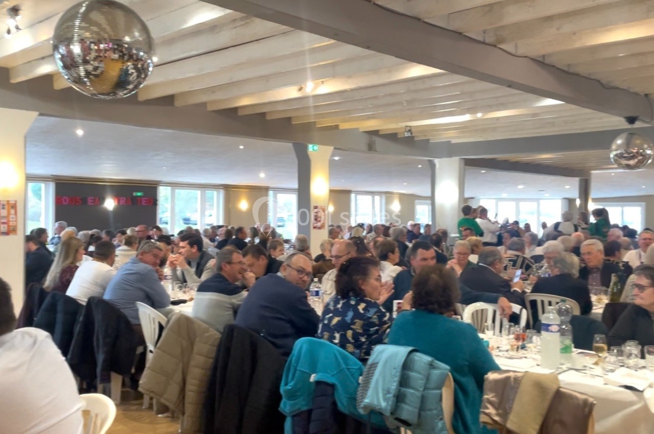 Salle remplie de personnes assises à des tables lors d'un événement, sous un plafond avec poutres apparentes et boule disco.