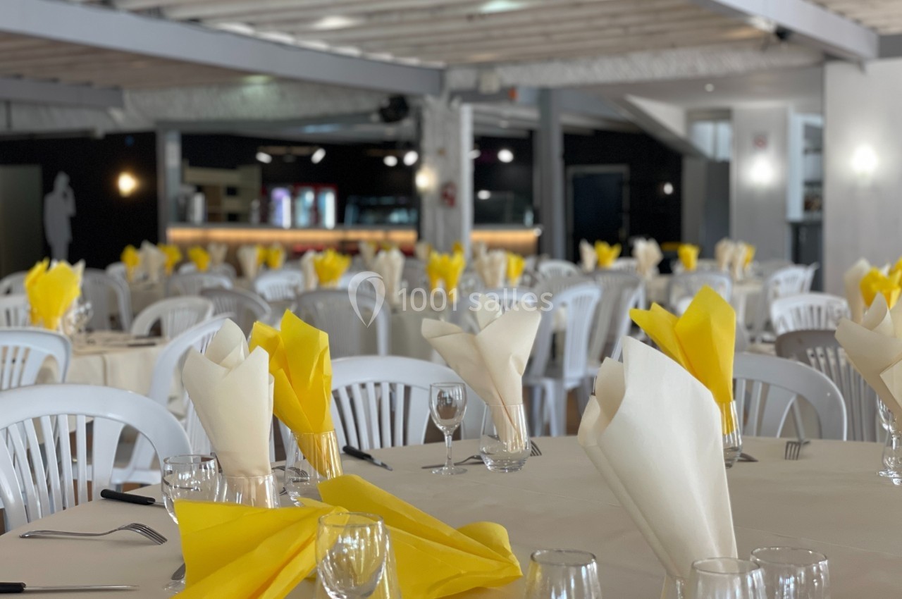 Salle de réception avec tables dressées, nappes blanches, serviettes jaunes et blanches, et chaises en plastique.